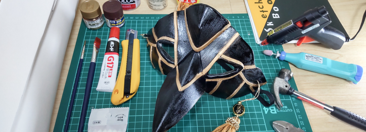 黒い烏天狗のお面の周りに接着剤やカッター、コスボードなどが置かれた作業台